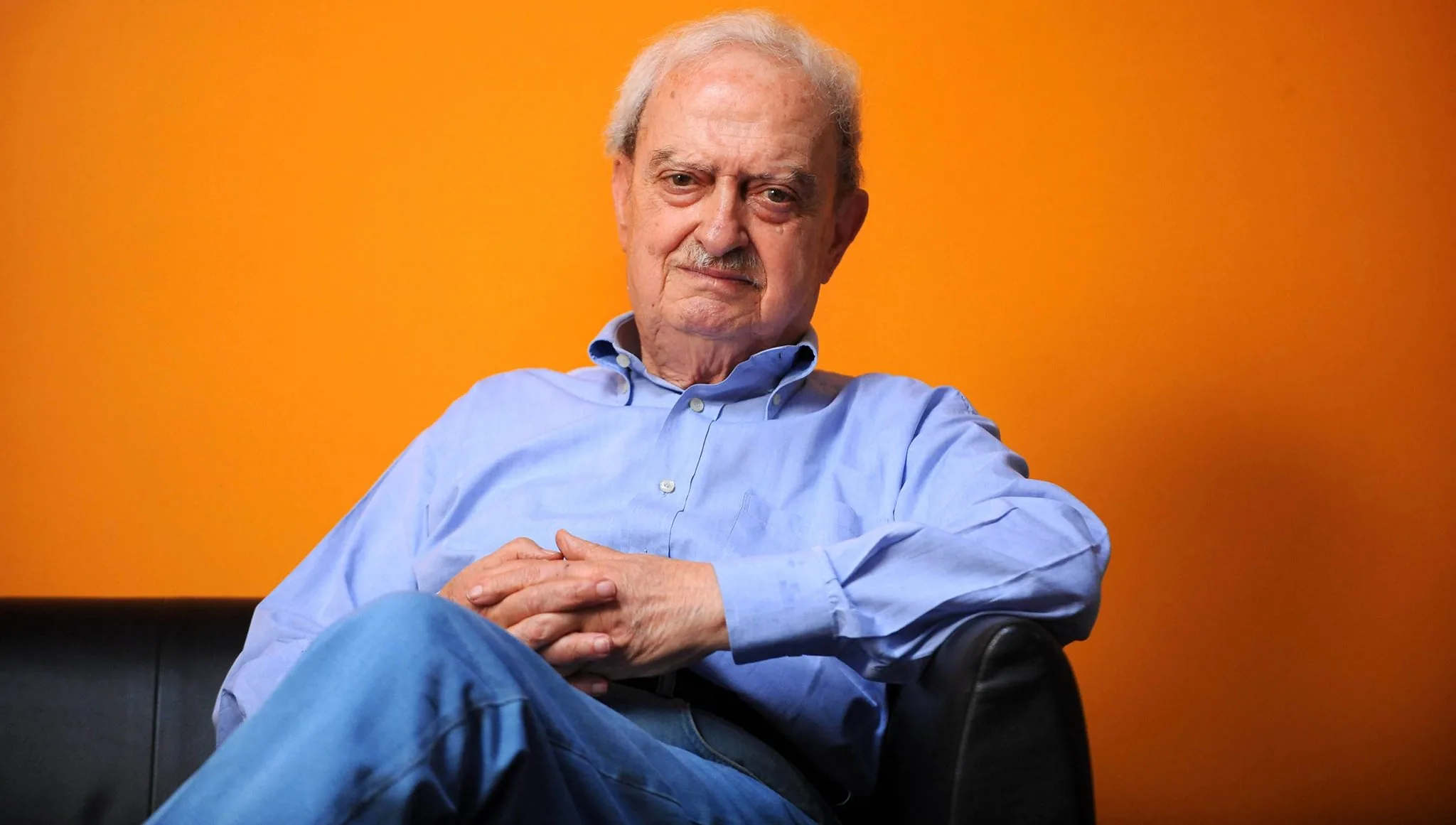 Emanuele Macaluso seduto su una poltrona, in primo piano, con sfondo arancione
