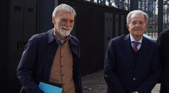 Due uomini anziani in abiti formali sorridono all’esterno durante un incontro pubblico o evento politico.