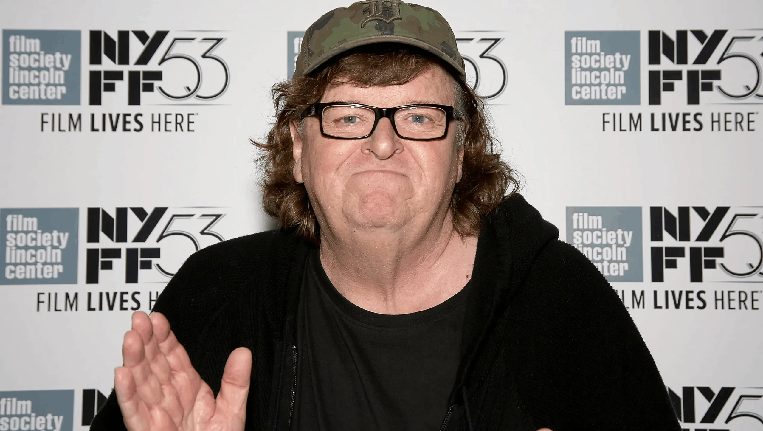 Michael Moore con cappellino e occhiali in posa durante un evento cinematografico, davanti a un backdrop con loghi del New York Film Festival.