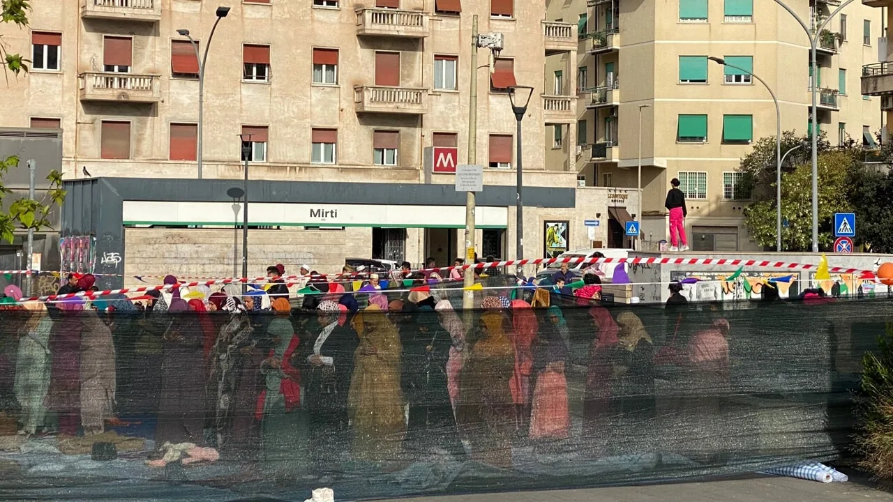 Donne migranti in fila davanti a un centro servizi in città, viste attraverso una rete di cantiere; sullo sfondo edifici popolari.