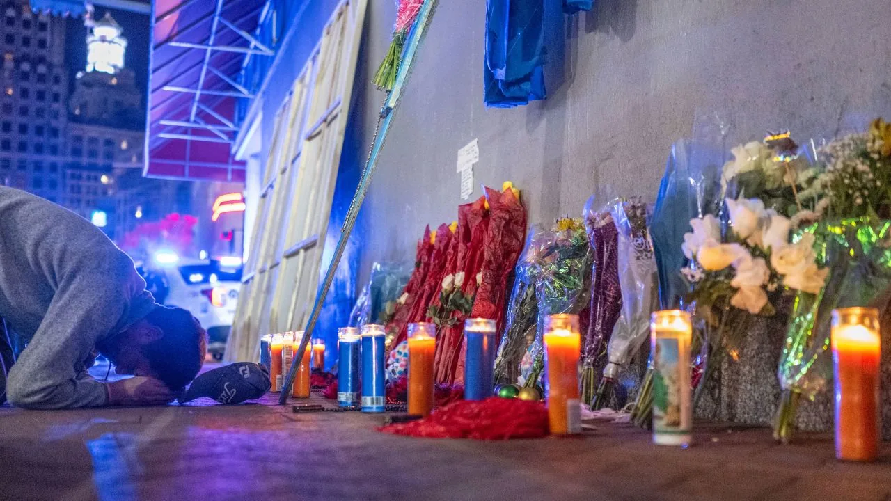 Uomo inginocchiato accanto a un memoriale con candele accese, fiori e luci blu, in ricordo delle vittime di un attacco.