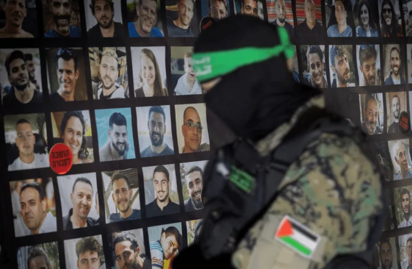 Uomo in uniforme militare con volto coperto davanti a una parete con fotografie di persone.