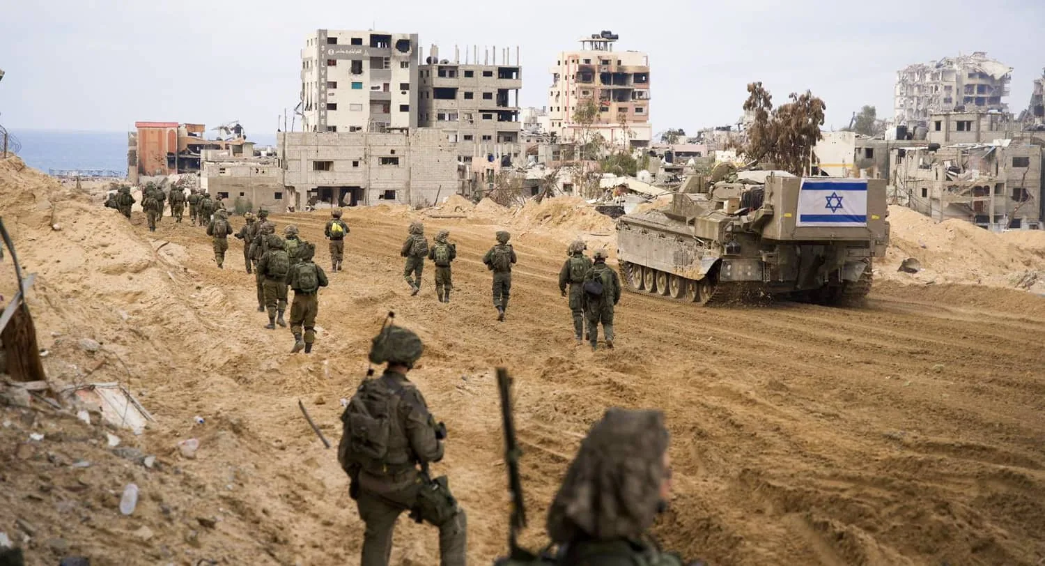 Soldati israeliani avanzano in una zona urbana distrutta durante un’operazione militare, con un carro armato e bandiera visibile.