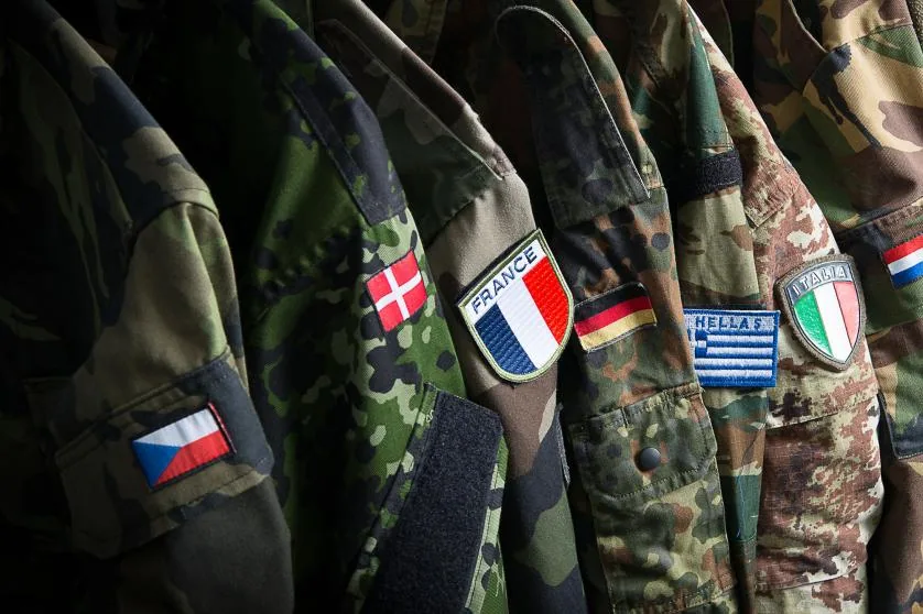 Uniformi militari di diversi paesi europei con bandiere nazionali cucite sulle maniche, simbolo della cooperazione nella difesa europea.